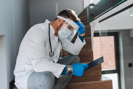 A Tired And Exhausted Doctor With Protective Face Mask And Shield Sitting On The Stairs In Hallway And Reading The Patient's Results. Pandemic, Covid-19, Epidemic, Coronavirus Concept.