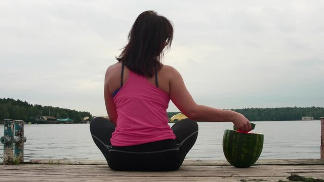Back view of woman in pibk tank sitting on old pier with watermelon behind her. She takes a slice of watermelon and eats it. Summer food.