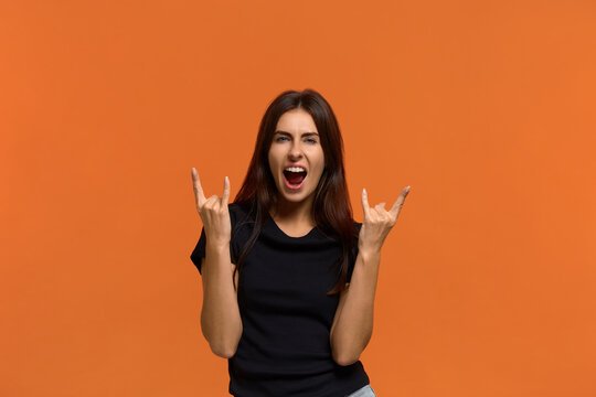 Lets have fun. Emotive caucasian woman in black t-shirt makes rock signs, feels self confident, yells loudly, ready for the party. Isolated over an orange background