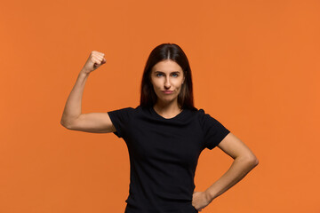 Girls power. Serious caucasian woman in black t-shirt raising hand while showing muscle, trying to look dangerous while. Ready to revenge everyone for bad things. Isolated over an orange background