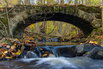 Fototapeta na wymiar A Water cascade in the creek flowing under an ancient stone bridge in the autumn forest.
