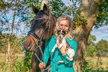 Beautiful portrait of a blond smiling girl with her horse and dog in the forest. Wearing a green...