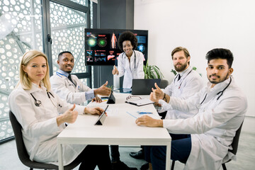 Team of smiling multiethnic doctors, Caucasian, African and Indian, having brainstorming session in the meeting room, sitting at the table and showing thumbs up to camera