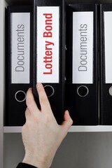 Lottery Bond. File Folder is taking by a hand from office shelf. Red Text is on the label of the...