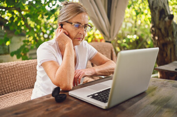 Happy senior woman use wireless headphones working online with laptop computer outdoor in garden