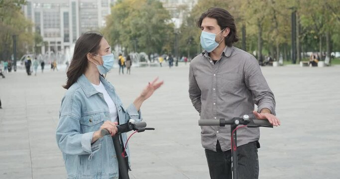 Couple wearing protective face mask and talking while leaning on e scooters in city