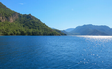 The green hill with trees is surrounded by blue still sea. Reflection of sunlights over the sea on a blue sky.