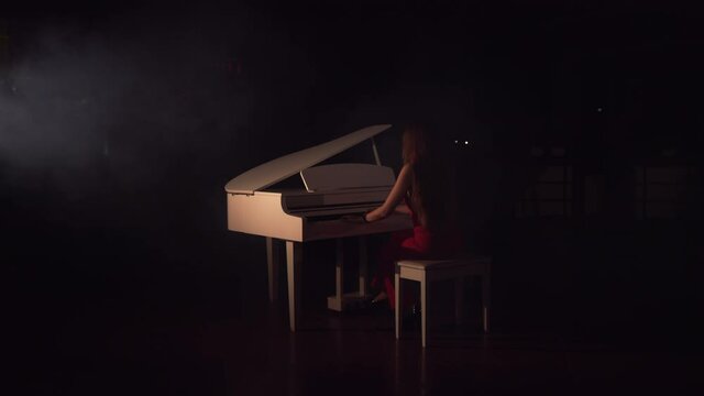 Pianist plays beautiful white grand piano on stage in concert . View from the back.Young beautiful girl in a red dress and plays the piano in a black room with a slight feeling of smoke