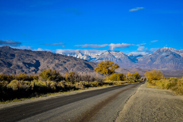 rural road to mountains with autumn trees and snow clouds in blue sky landscape