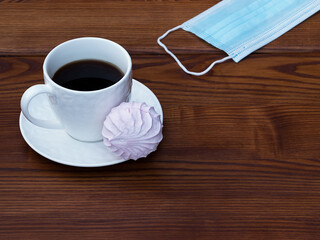 Obraz premium White cup of coffee and pink zefir on the wooden background. Traditional russian sweets like marshmallow. Copy space. Medical mask on the side.