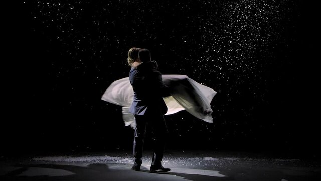 Tender couple in love on the background of falling snow. A man is circling in his arms his beloved woman on a black background in the rays of studio light. Love story, first snow, romance. Slow motion