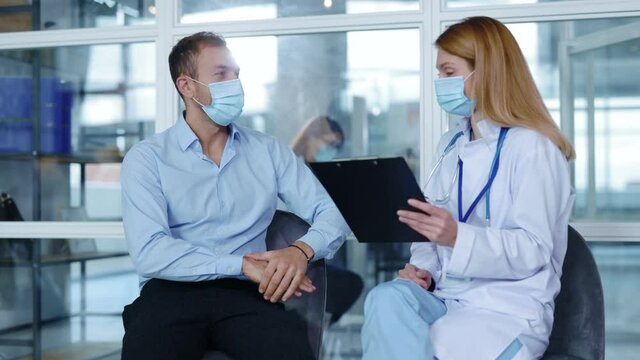 Portrait attractive man with medical mask sitting speaking with woman doctor about diagnosis during consultation. Patient clinic covid coronavirus illness. Slow motion