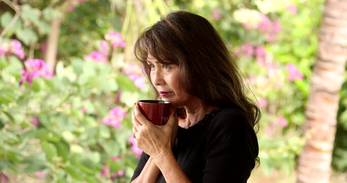 Attractive asian woman with long dark hair and wearing black top drinking coffee in medium shot on a lanai. Bright background, with tropical green and purple flowers.