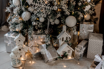 Christmas, New Year interior decorated Christmas gifts with ribbons, white figures of  houses , basket with wood and Christmas tree with white and silver balls,  snowflakes, bump and  glowing garland.