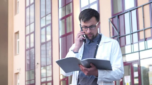Male doctor in white lab coat talking on the phone with patient while examining his medical records outside the clinic. Physician consulting patient on test results. Personally delivering feedback
