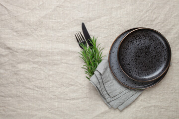 Black plate, cutlery and napkin with a sprig of rosemary on textile table. Top view. Table setting.