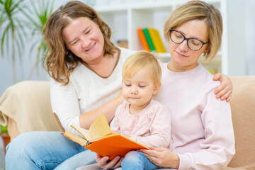 Obraz premium Two young women are sitting in the living room on the couch with a child on their knees and are reading a book to the book. Gay lesbian couple at home with a baby. Lesbian family concept