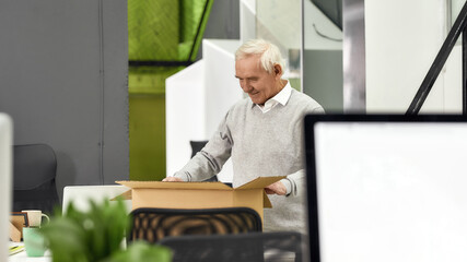New employee aged man, senior intern looking happy while unpacking box with personal belongings, preparing for first day at work in the office