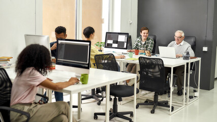 Full length shot of aged man, senior intern typing, using laptop while sitting at desk, working together with other young employees in modern office