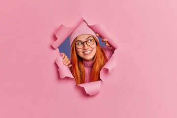 Curious cheerful redhead woman concentrated aside sees something very pleasant smiles broadly wears hat and spectacles poses in ripped hole of pink background. Foxy happy teenage girl at studio