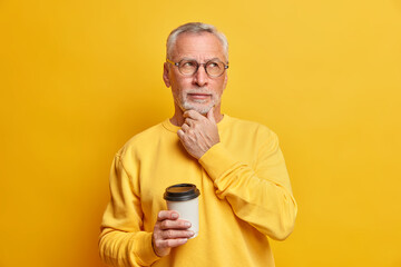 Pensive bearded male pensioner holds chin and concentrated aside drinks takeaway coffee and has...