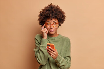 Fototapeta na wymiar Pensive dark skinned young ethnic woman keeps index finger on temple tries to remember telephone number of someone holds modern smartphone and concentrated aside wears glasses casual jumper.