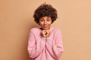 Good looking woman with curly hair keeps hands under chin looks direcly at camera listens something attentively wears casual knitted sweater isolated over brown background admires wonderful thing