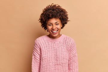 Fototapeta na wymiar Beautiful sincere woman with Afro hair smiles broadly has white perfect teeth doesnt hide positive emotions wears knitted sweater isolated over brown background. Glad optimistic female model
