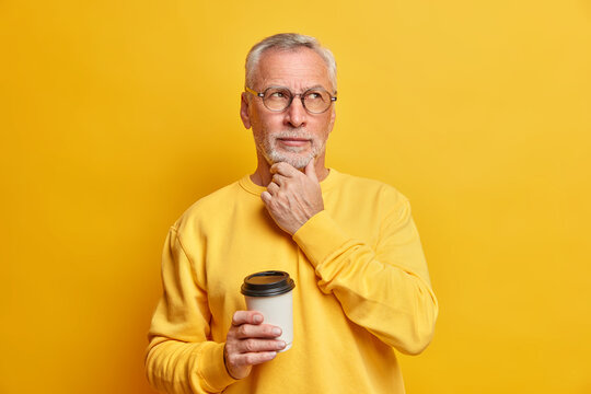 Pensive bearded male pensioner holds chin and concentrated aside drinks takeaway coffee and has break dressed in casual jumper isolated over yellow background. Thoughtful grandfather indoor.