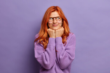 Pleased redhead woman keeps hands under chin concentrated aside has dreamy expression wears optical glasses and purple sweater poses indoor has intention to do something. Thoughtful ginger female