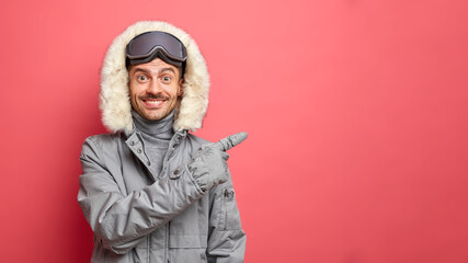 Handsome smiling male snowboarder in warm winter clothing has happy expression points on right at empty space isolated over bright pink background enjoys adventure and travel. Holiday concept