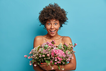 Greatly surprised dark skinned woman poses with bare shoulders holds big bunch of flowers gets bouquet from husband has pleased face expression isolated over blue background. Celebration concept