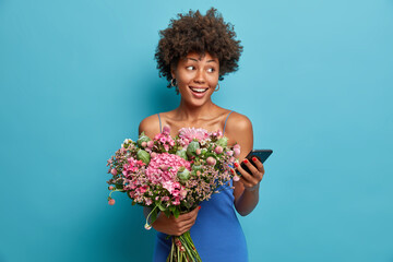 Positive dark skinned woman with curly hair holds nice bouquet of flowers gets order online uses modern smartphone enjoys present from boyfriend looks aside with smile isolated on blue wall. - Powered by Adobe