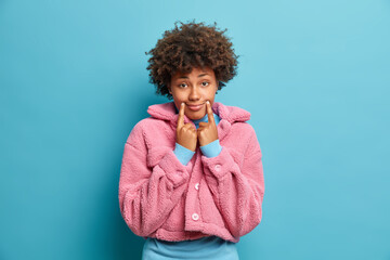 Sad dark skinned woman tries to be happy keeps finger near corners of lips forces smile looks with gloomy expression at camera wears pink fur coat isolated over blue background. Negative emotions