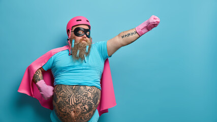 Self confident serious bearded man ready to help people wears superhero costume pretends being heroic character has supernatural power stretches arm over blue background with empty blank space - obrazy, fototapety, plakaty
