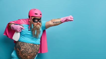 Horizontal shot of self confident male hero wears pink helmet cloak and rubber gloves holds bottle of detergent ready to help you with cleaning stretches arm as going to fly isolated over blue wall - obrazy, fototapety, plakaty