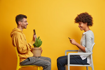 Interracial couple use modern gadgets sit opposite each other on chairs spend free time at home holds potted cactus isolated over yellow background use high speed internet. Technology concept