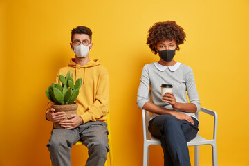 Horizontal shot of mixed race woman and man wear face masks to prevent from corona virus pose on seperate chairs keep social distancing isolated on yellow background dont go outside during quarantine