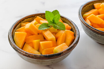 Cantaloupe melon bite-sized cubes served with mint leaves close up in a bowls on light marble background. Fruit salad, dessert