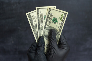 A man's hand in black medical gloves holds three hundred dollar bills in his hand