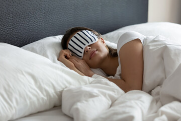 Stylish nightwear. Comfy sleeping mask helping young woman tourist traveler enjoy good healthy night sleep at new place in hotel suite room, millennial woman napping at wide king size bed in eye mask - obrazy, fototapety, plakaty
