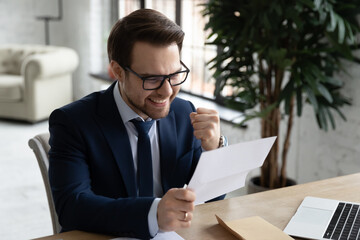 Overjoyed young male manager in formal suit and glasses reading unexpected win news paper...