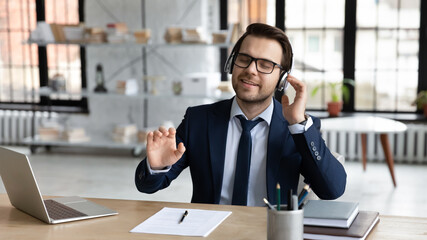 Relaxed young calm handsome smiling businessman manager in formal wear and eyeglasses sitting at workplace, enjoying stress free pause time listening favorite music tracks in modern earphones.