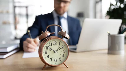 Foto op Plexiglas Focus on clock standing on table with busy young businessman entrepreneur in formal wear on background, focused millennial male manager involved in casual working process, time management concept. © fizkes