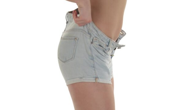 Side view of unrecognizable slender Caucasian woman getting dressed. Profile view of model wearing and zipping tight denim shorts. Isolated on white background.