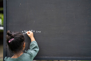 Cute little girl writing alphabet on blackboard in garden.
