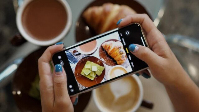 Close up of woman holds modern smartphone and taking pictures of her morning breakfast coffee, cocoa and tasty croissant buns wit green chocolate to share photos on social media resources