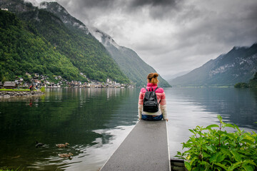 The girl sits on the bridge and looks at the foggy autumn town. Hallstatt, Austria. Girl with a backpack back