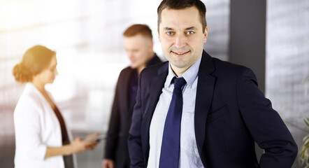 Portrait of a self-confident middle aged businessman in a blue suit, standing in a sunny modern office with his colleagues at the background. Concept of business success
