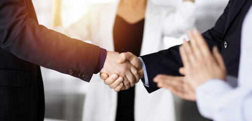 Unknown businesspeople are shaking their hands after signing a contract, while standing together in a sunny modern office, close-up. Business communication, handshake, and marketing concept